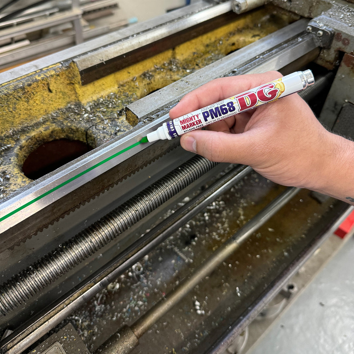 A hand holding a DG Mighty Marker PM68 green Mighty Marker drawing a green line on a piece of metal machinery inside a workshop.