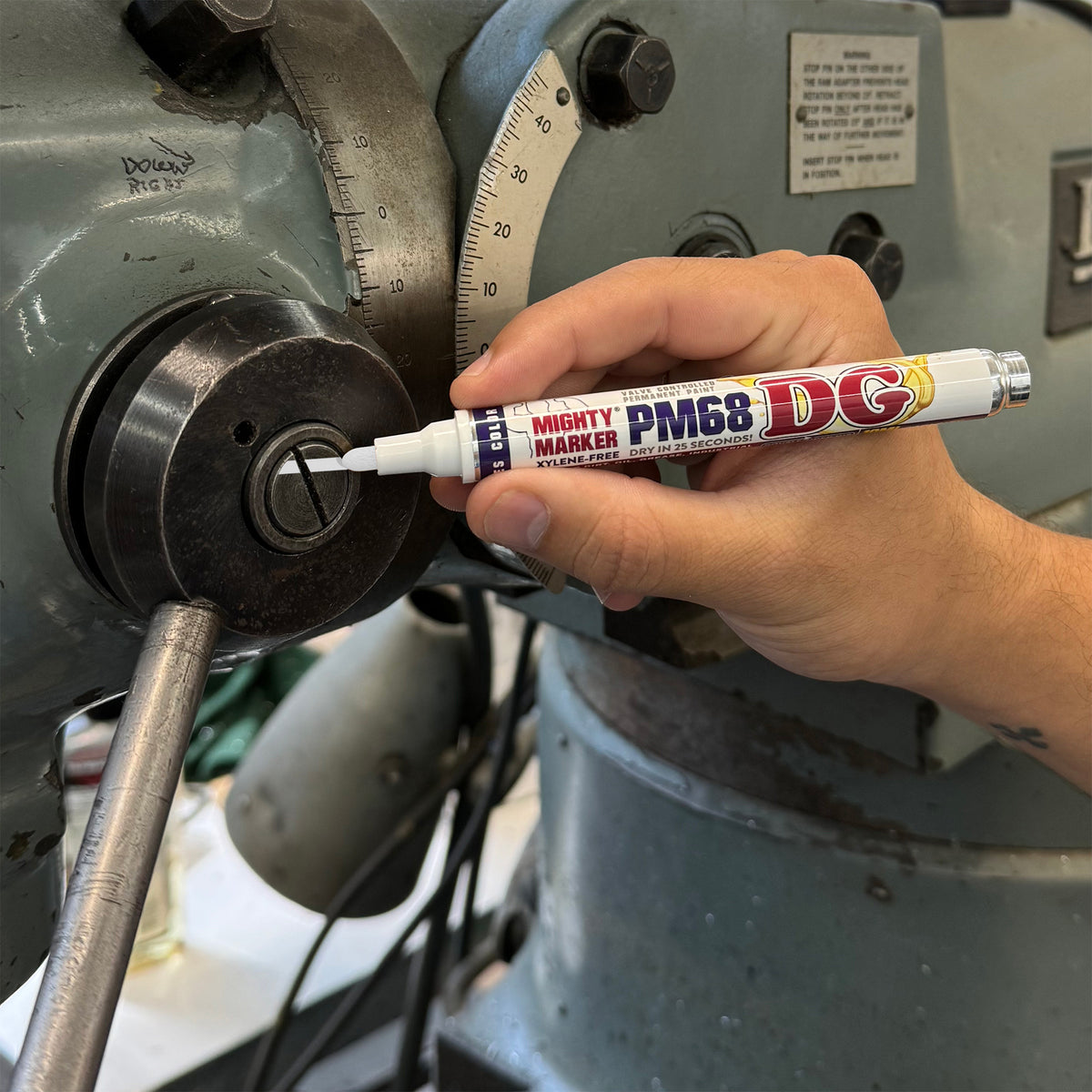 A hand holding a white Mighty Marker PM68 DG marking a circular dial on a large gray industrial machine.