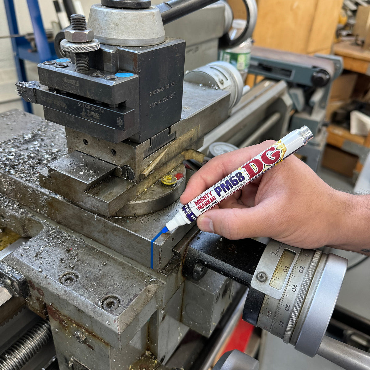 A hand holding a Blue DG PM68 Mighty Marker applies a blue marking line to a machined metal surface on a lathe machine in a workshop setting.